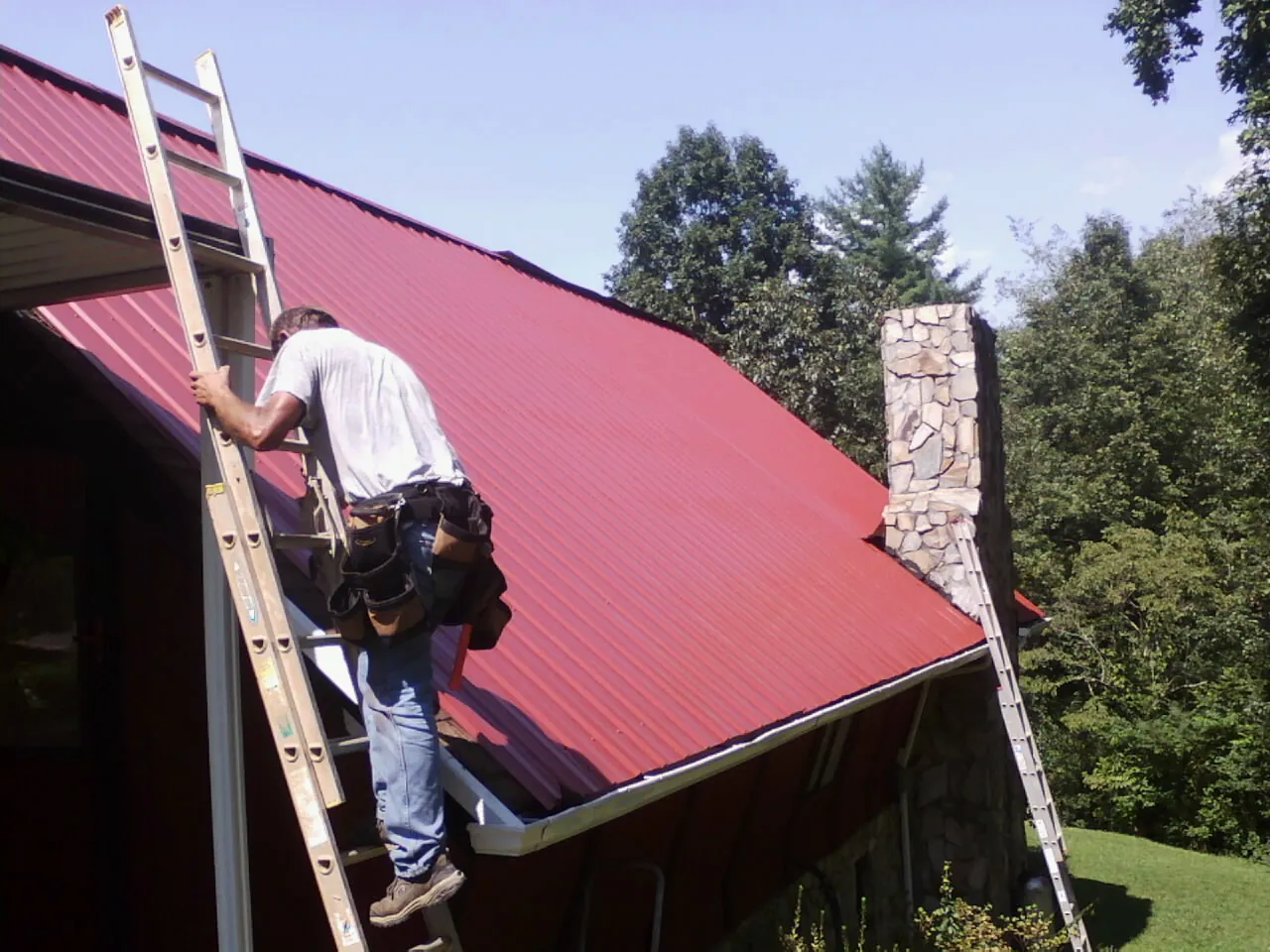 Roofer on a ladder beside a red metal roof and stone chimney — Soffit & Fascia Repair by Roofing Brothers in Swarthmore